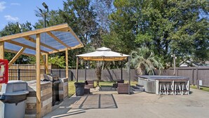 Un patio exterior con zona de parrilla cubierta, una mesa con sillas y un cubo de basura.