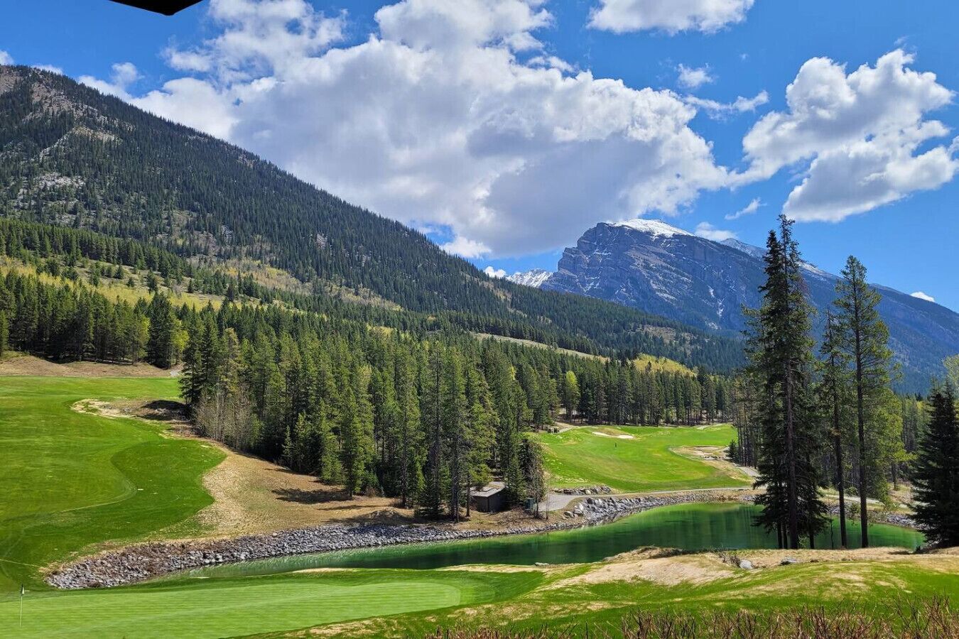 A golf course with a pond, surrounded by dense forests and mountains.