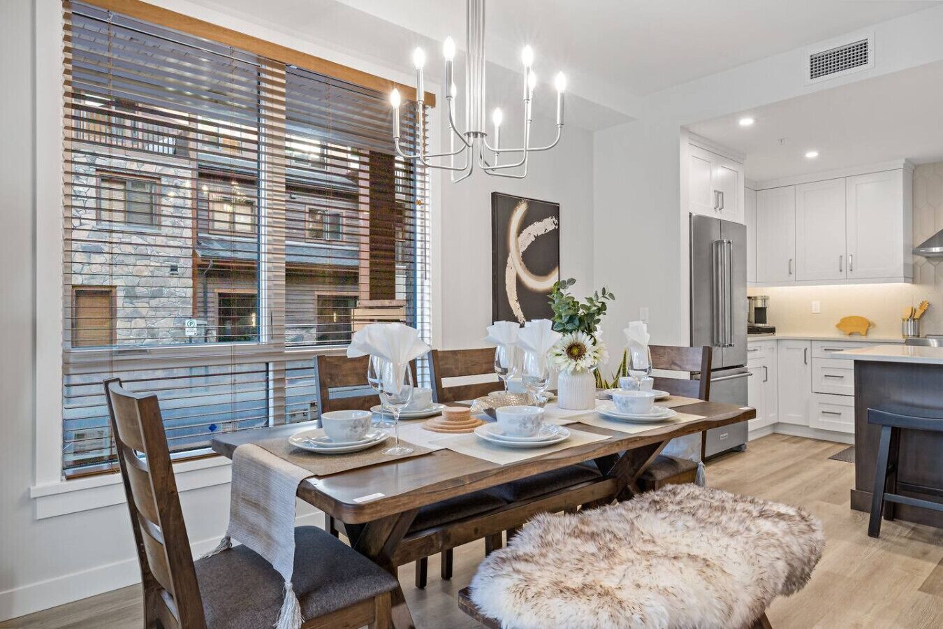 Un espace de salle à manger moderne avec une table en bois dressée pour quatre personnes, un lustre et une grande fenêtre équipée de stores.