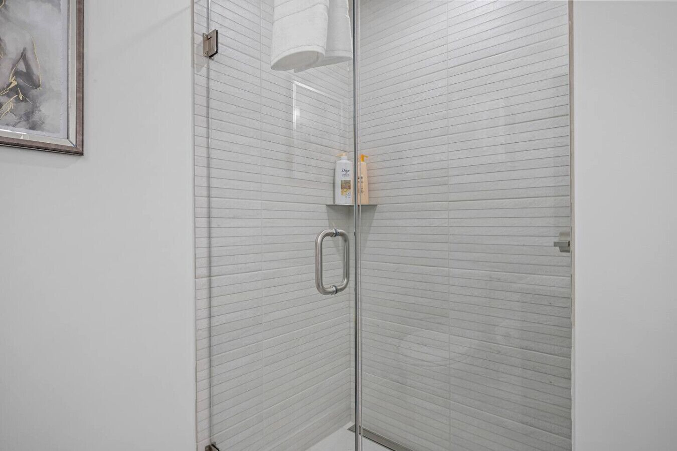 Une douche fermée par des parois de verre, dotée d’une poignée en métal, de murs recouverts de carreaux blancs et d’un tableau encadré sur le côté gauche.