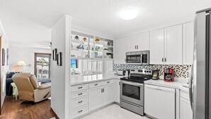 Eine moderne Küche mit weißen Schränken, einer Mikrowelle, einem Backofen und einem Geschirrspüler. Auf der Arbeitsplatte steht eine Kaffeemaschine und in der Ecke ein Stuhl.