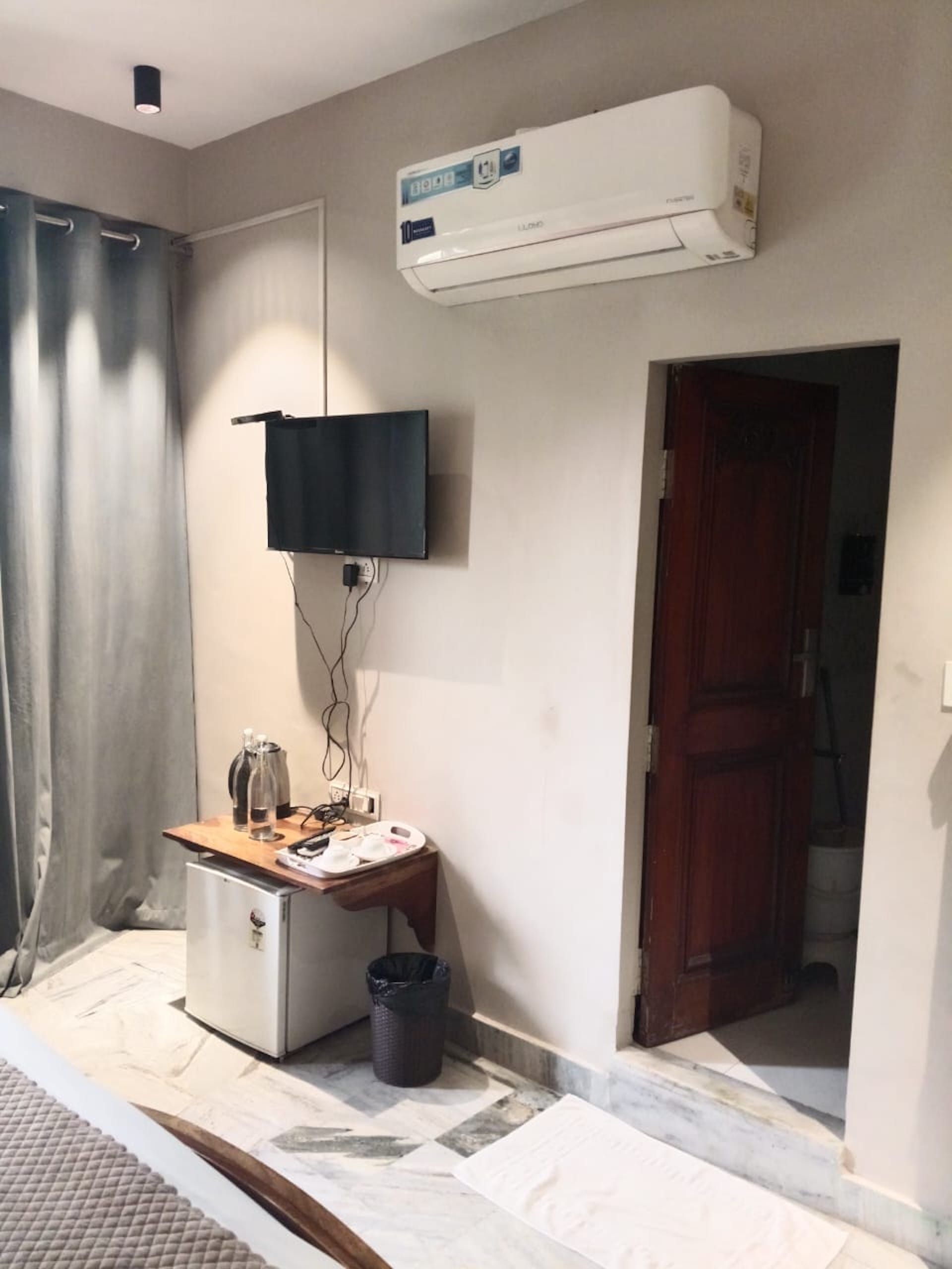 A hotel room with a flat-screen TV mounted on the wall, a small table with a kettle and a trash bin, and an air conditioning unit above the table.