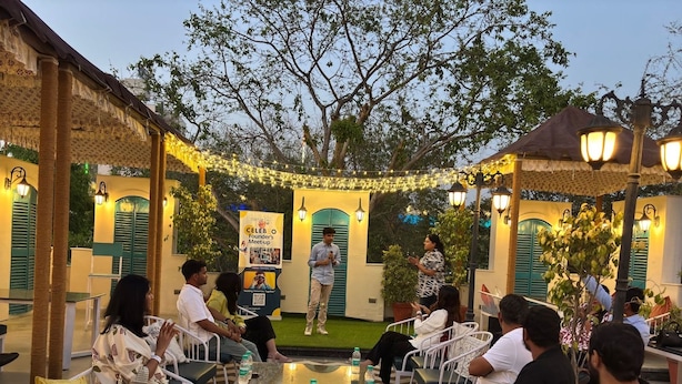 An outdoor event with attendees seated at tables, a speaker addressing the audience, and decorative string lights. - J Startup House (Jaipur)