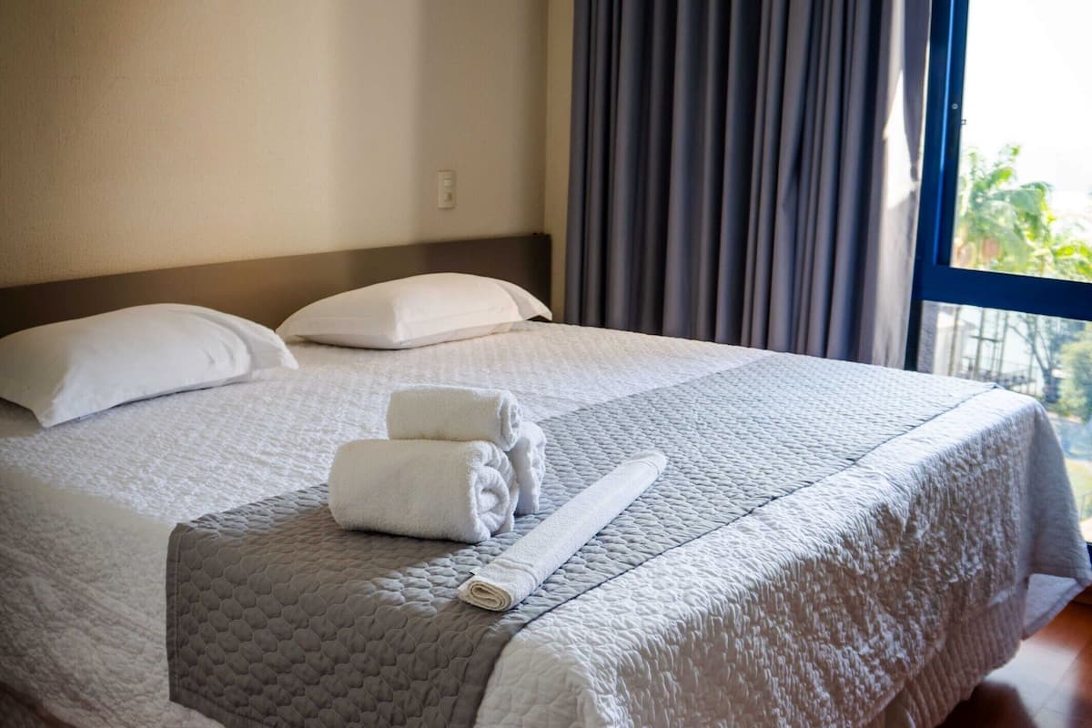 Un lit bien fait, avec du linge de lit blanc et une couverture grise, une fenêtre avec des rideaux et un plancher en bois.
