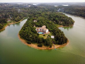 Luchtbeeld van een afgelegen eiland met een groot gebouw, inclusief een zwembad, omgeven door dicht bos en een kalm meer.