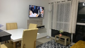 Un salon avec un téléviseur fixé au mur, une table à manger avec des chaises, une petite table d’appoint et une fenêtre avec des rideaux.