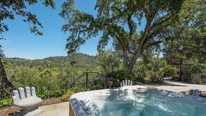 Un jacuzzi con un banco con forma de mano de piedra, rodeado de abundante vegetación y un cielo azul despejado.