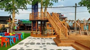 Una estructura de juego de madera con una escalera, rodeada de sillas y mesas de plástico de colores sobre un suelo con diseño de guijarros.