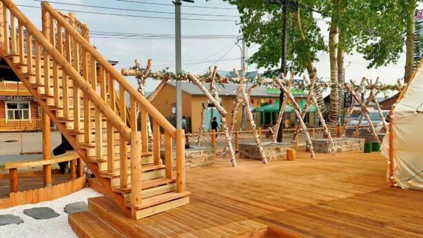 Una escalera de madera que conduce a un espacio cubierto con piso de baldosas, rodeado de árboles y edificios.