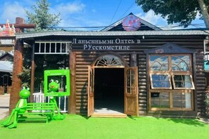 Un edificio de madera con un letrero que dice 'Ляпузовский Отель в РУССКОМТЫЕ' y una estatua de rana verde en el exterior.