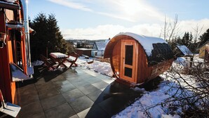 Een houten blokhut met een sneeuwbedekt dak, een terras met een tafel en stoelen, en een uitzicht op het omliggende landschap.