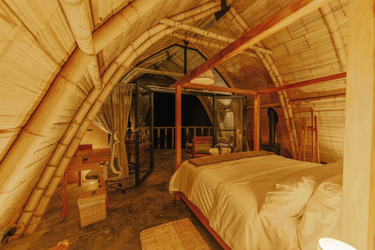 A room with a sloped bamboo roof, a bed with white bedding, a wooden chair, and a small table.