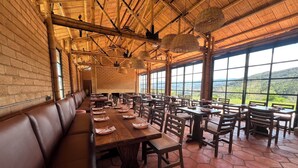 A restaurant with a long wooden bench, tables, and chairs. Large windows offer a view of the outdoors. - Hotel Casa Selvaggio (Ráquira)