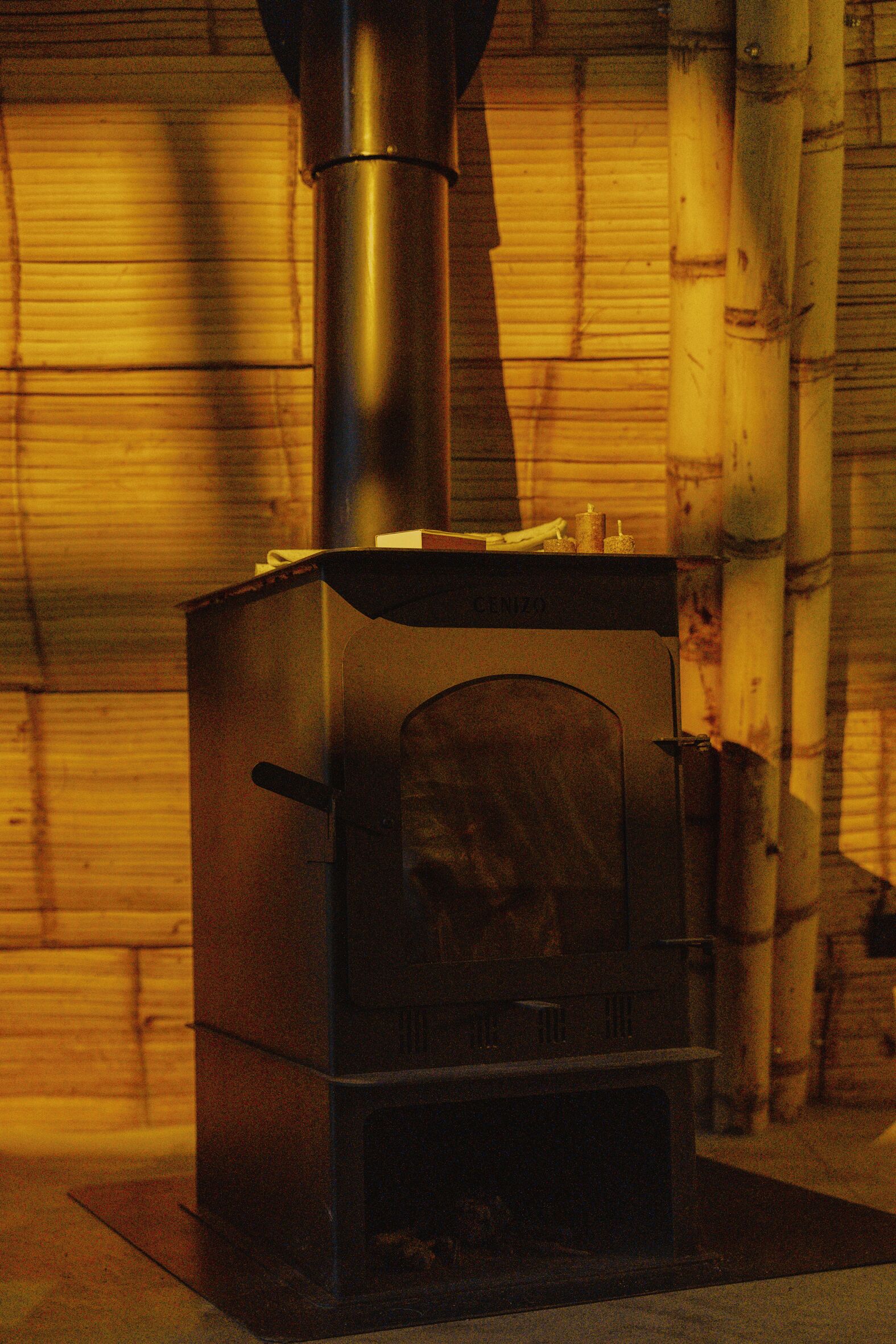A wood-burning stove with a chimney, placed in a room with wooden walls and a bamboo structure to the right.