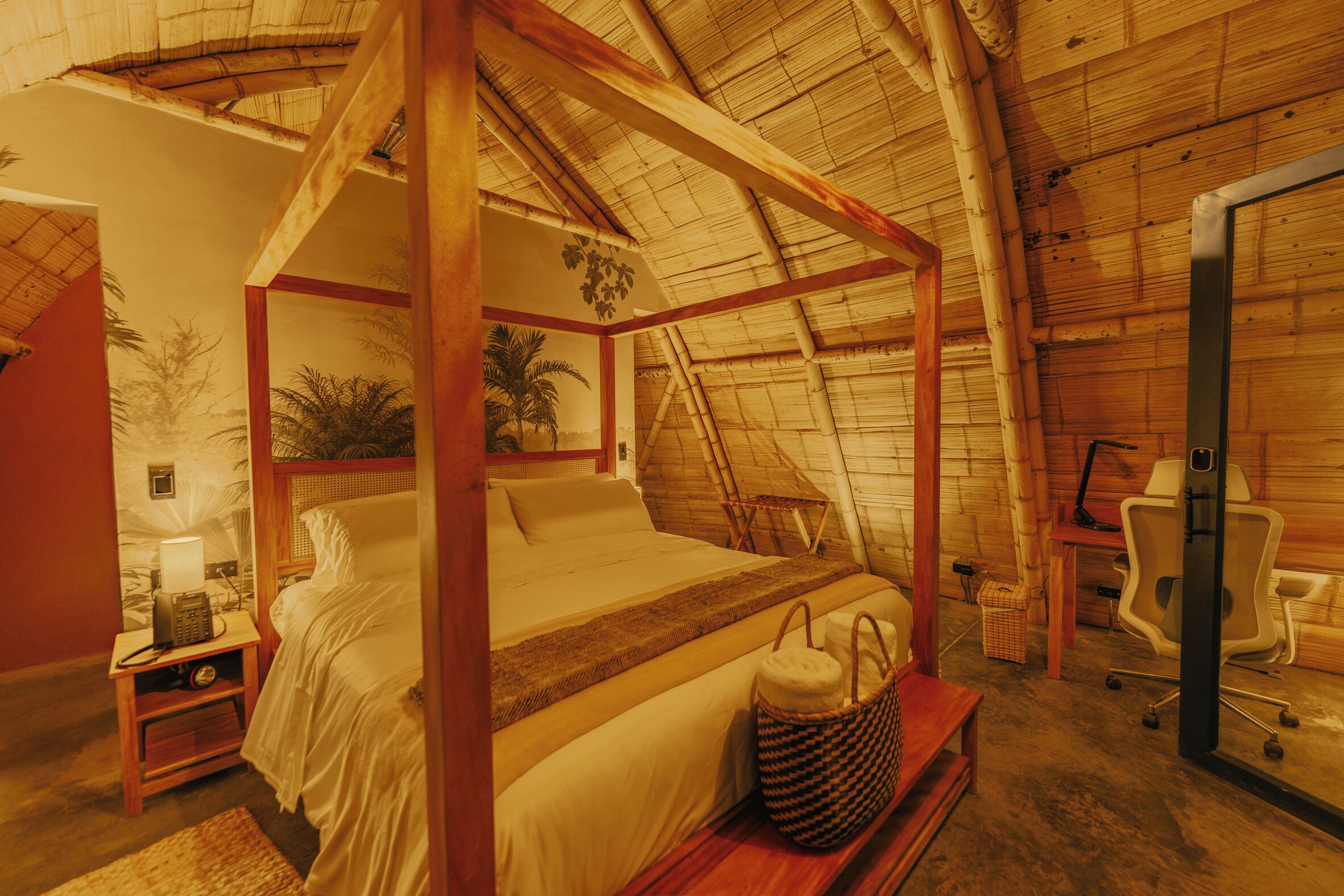 A room with a four-poster bed, a desk with a chair, a lamp, and a small table with a basket.