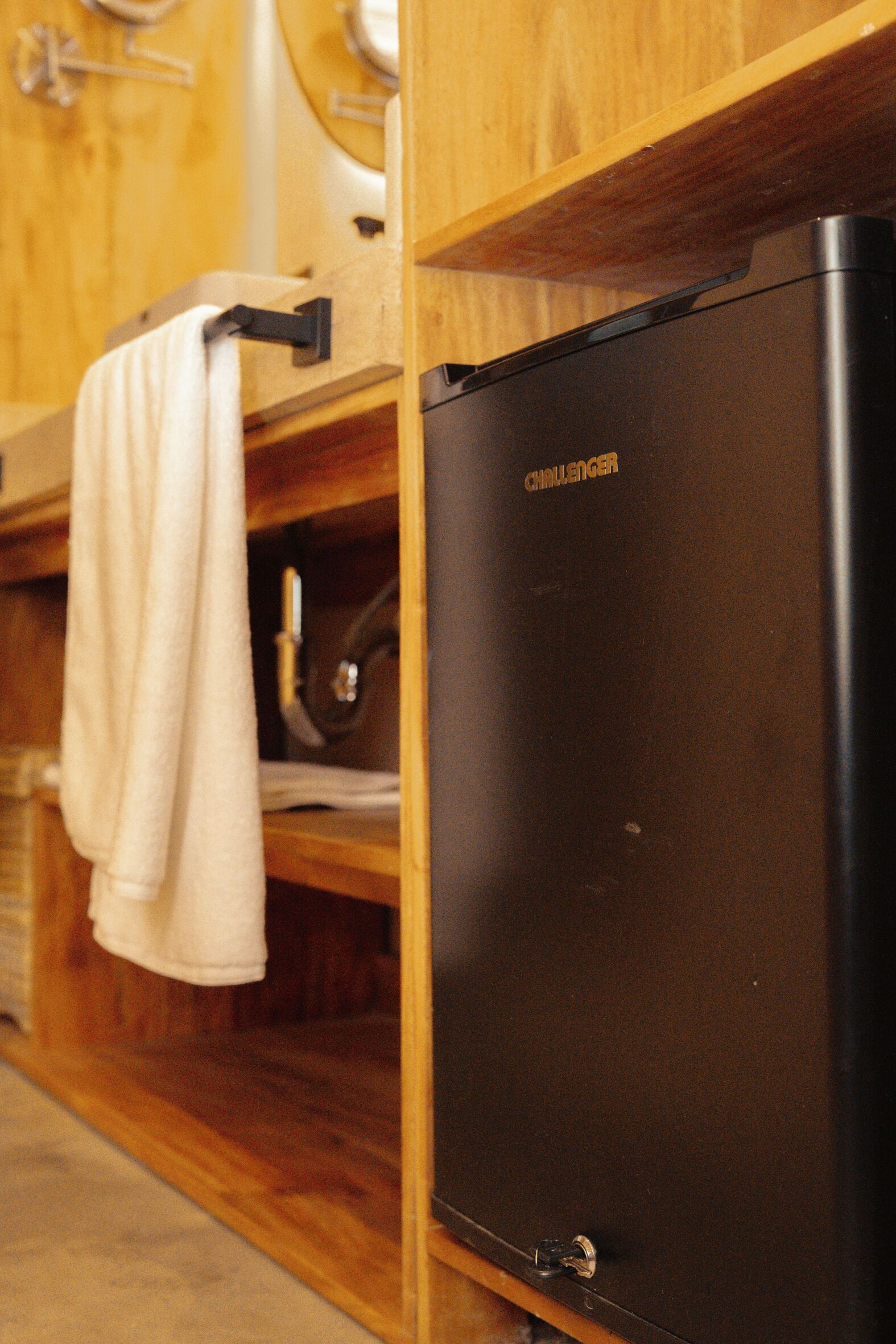 A compact black refrigerator with the brand name "CHALLENGER" in gold lettering, a wooden cabinet with a towel hanging on a metal hook, and a wooden countertop.