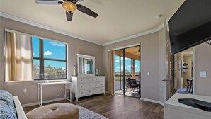 Une chambre avec un lit, une commode, un bureau, une chaise, un ventilateur de plafond et un balcon avec une table et des chaises.