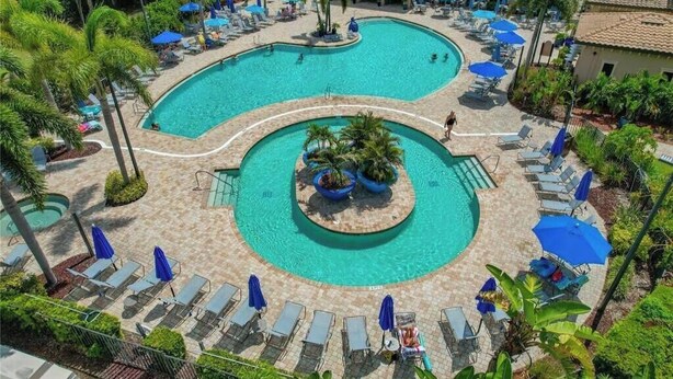 Une grande piscine à plusieurs niveaux, dotée de chaises longues et de parasols, entourée de palmiers et d’un bâtiment.