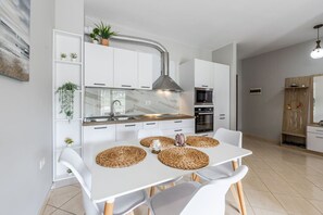 Une cuisine moderne avec des armoires blanches, un évier en acier inoxydable et un four encastré. On y trouve une table à manger avec des sets de table en fibres naturelles et des chaises blanches.