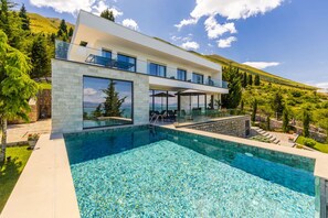 Ein modernes, zweistöckiges Haus mit großem Schwimmbecken, Glasbalkon und Blick auf die umliegende Landschaft.