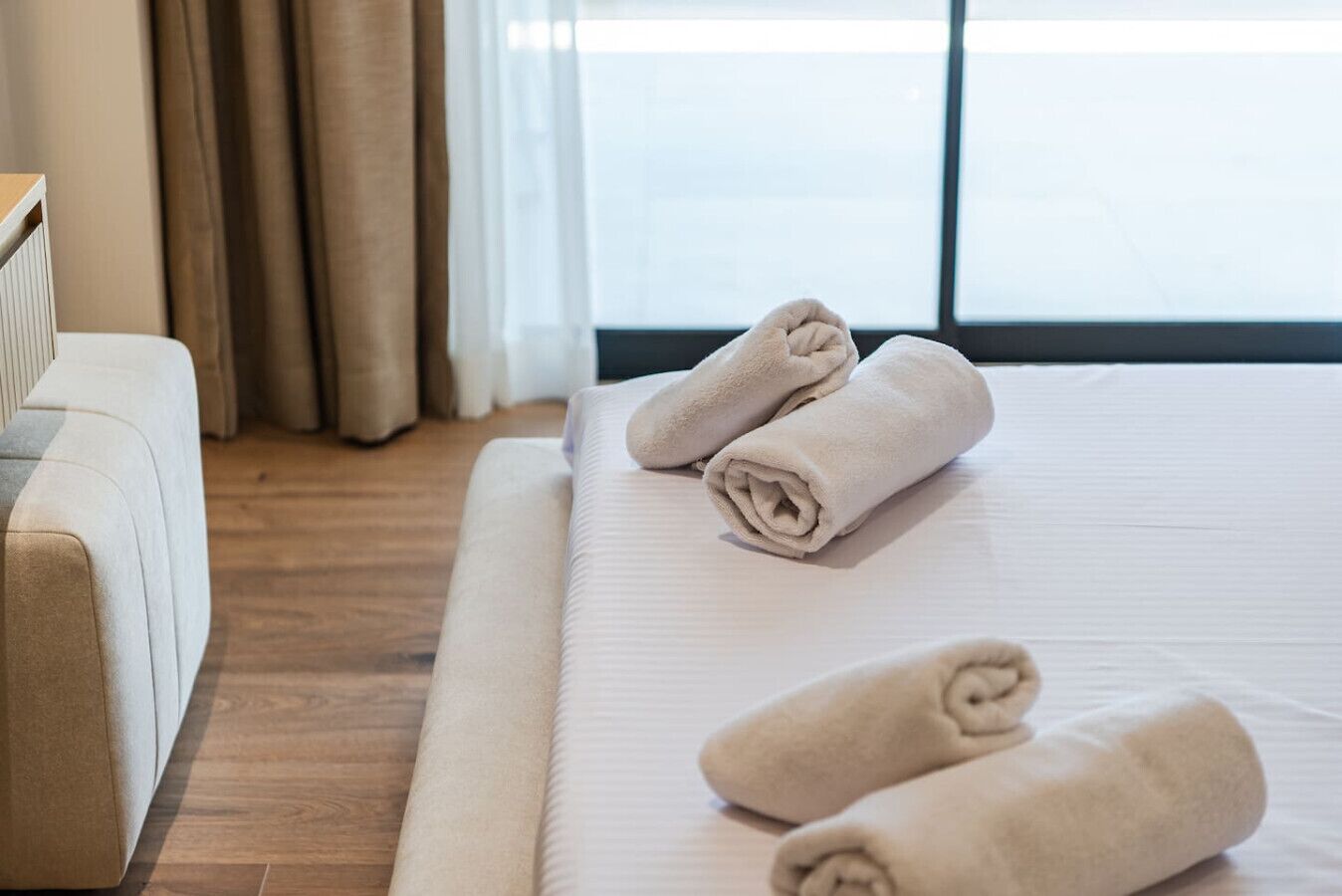 Ein Hotelzimmer mit einem Bett, zwei gerollten Handtüchern, Holzboden und einem Fenster mit Vorhängen.