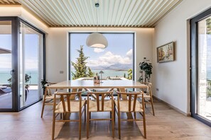 Ein moderner Essbereich mit großem Tisch und Stühlen, einer Pendelleuchte und Blick auf das Meer und die Berge durch ein großes Fenster.