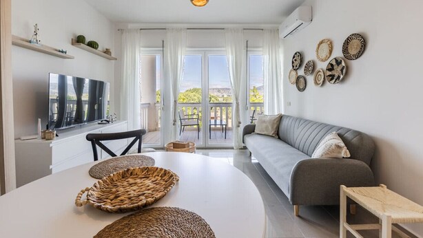 A modern living room with a grey sofa, a white dining table with woven baskets, a TV on the wall, and a balcony with a view. - Velipoja Beach Apartment by PikHost (Velipojë)