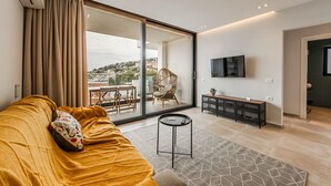 A modern living room with a sofa, a small table, a TV, and a sliding glass door leading to a balcony. - Ionian Sol Apartment | Blue Horizon Stay by PikHost (Sarandë)