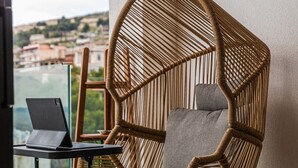 A wicker chair with a laptop on a small table, a view of a cityscape through a window, and a tiled floor. - Ionian Sol Apartment | Blue Horizon Stay by PikHost (Sarandë)