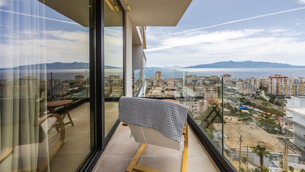 A balcony with a view of a coastal cityscape, a checkered table, and a chair. - Ionian Sol Apartment | Blue Horizon Stay by PikHost (Sarandë)