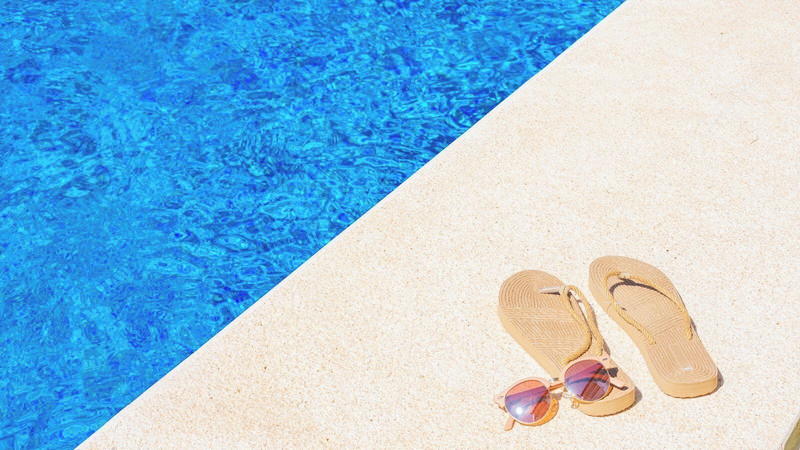 A pair of flip-flops and sunglasses on a sandy surface next to a pool with clear blue water.