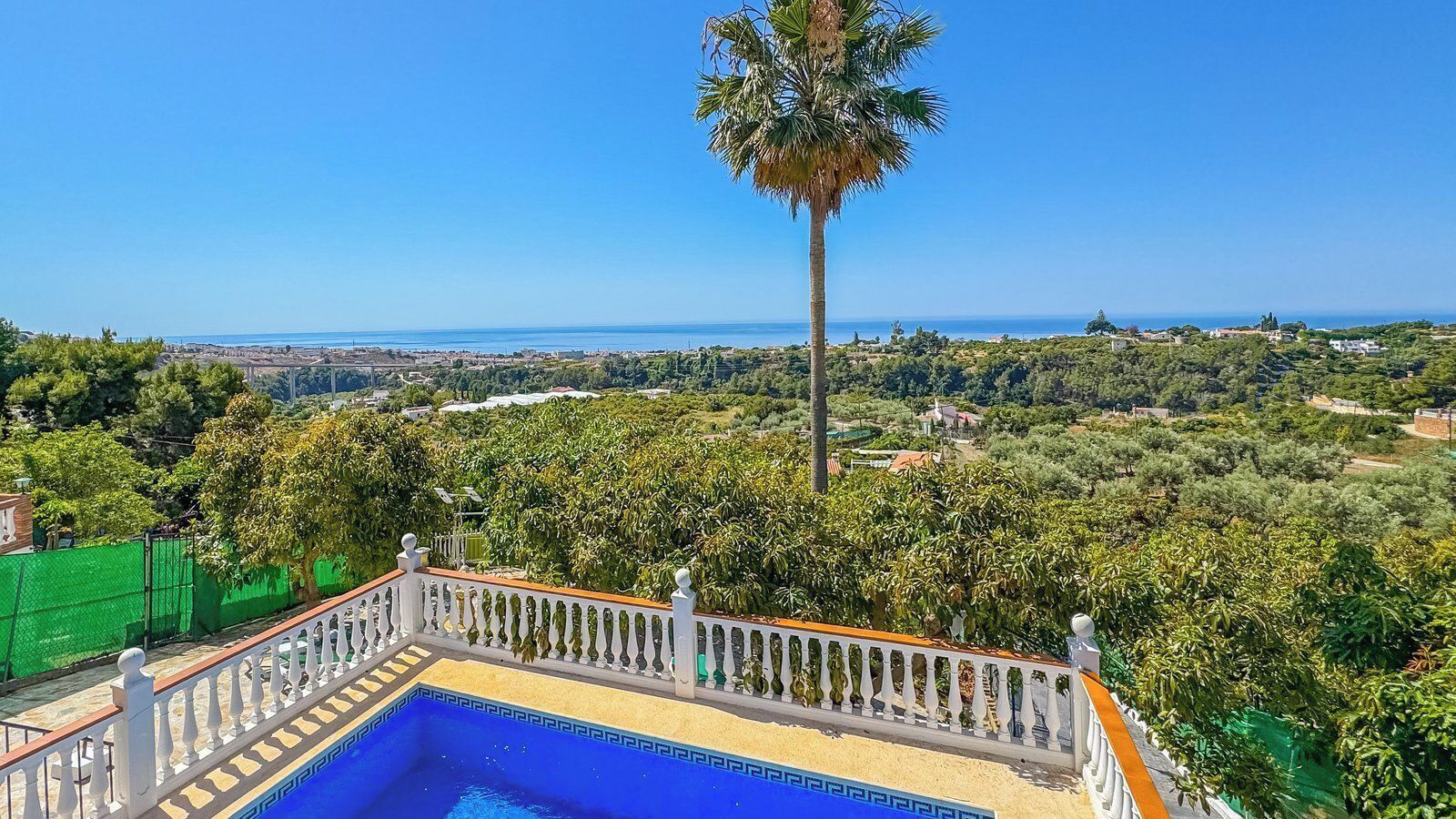 A swimming pool with a white balustrade, surrounded by lush greenery and a clear view of the coastline.