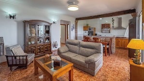 A living room with a sofa, armchair, and glass coffee table. There is a wooden cabinet with glass doors, a dining area with a table and chairs, and a kitchen with cabinets and appliances.