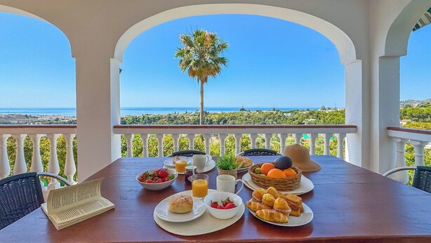 A dining table set with breakfast items, a book, and a view of the sea and greenery.