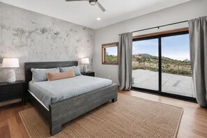 Een moderne slaapkamer met een groot bed, twee nachtkastjes, een plafondventilator en uitzicht op een landschap door een schuifdeur van glas.