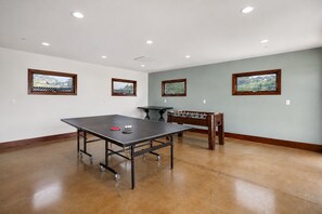 A spacious room with a ping pong table, a foosball table, and three windows with views of greenery.