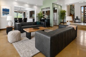 Een moderne woonkamer met een bank, salontafel en poef, een keuken op de achtergrond en een trap naar een hoger niveau.
