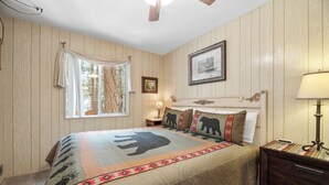 A bedroom with wooden panel walls, a bed with a bear-themed quilt, a bedside lamp, a framed picture on the wall, and a window with curtains. - Sky Krest (Big Bear Lake)