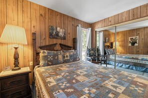 A bedroom with a wooden paneled wall, a quilted bedspread, a nightstand with a lamp, and a mirror reflecting the room. - Sky Krest (Big Bear Lake)