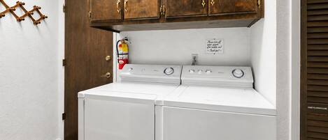 A laundry room with a white washer and dryer, wooden cabinets above, and a fire extinguisher on the wall.