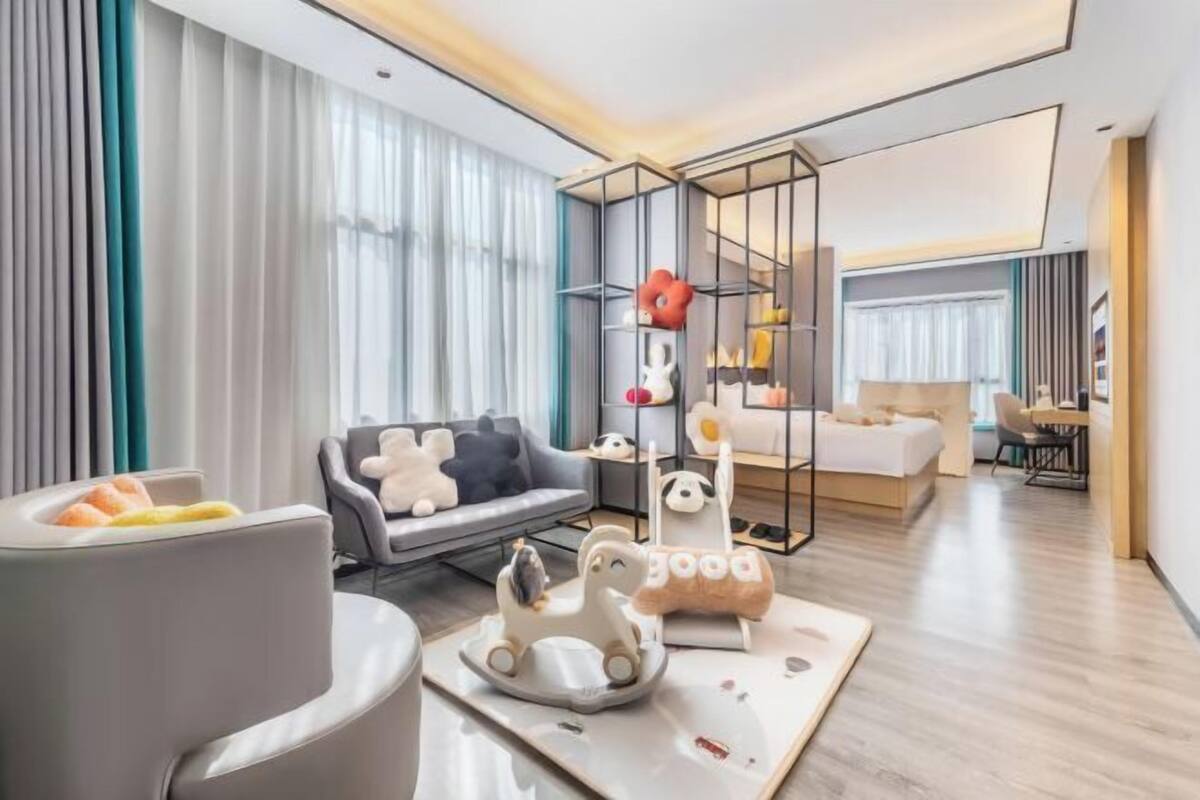 Moderni hotellihuone, jossa on harmaa sohva, sänky, työpöytä ja pieni leikkialue vauvoille.