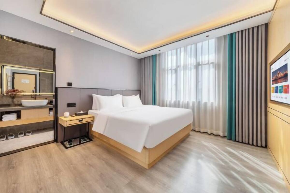 Moderni hotellihuone, jossa on suuri sänky, yöpöytä, sisäänrakennettu pesuallas ja suuri ikkuna, jossa on verhot.