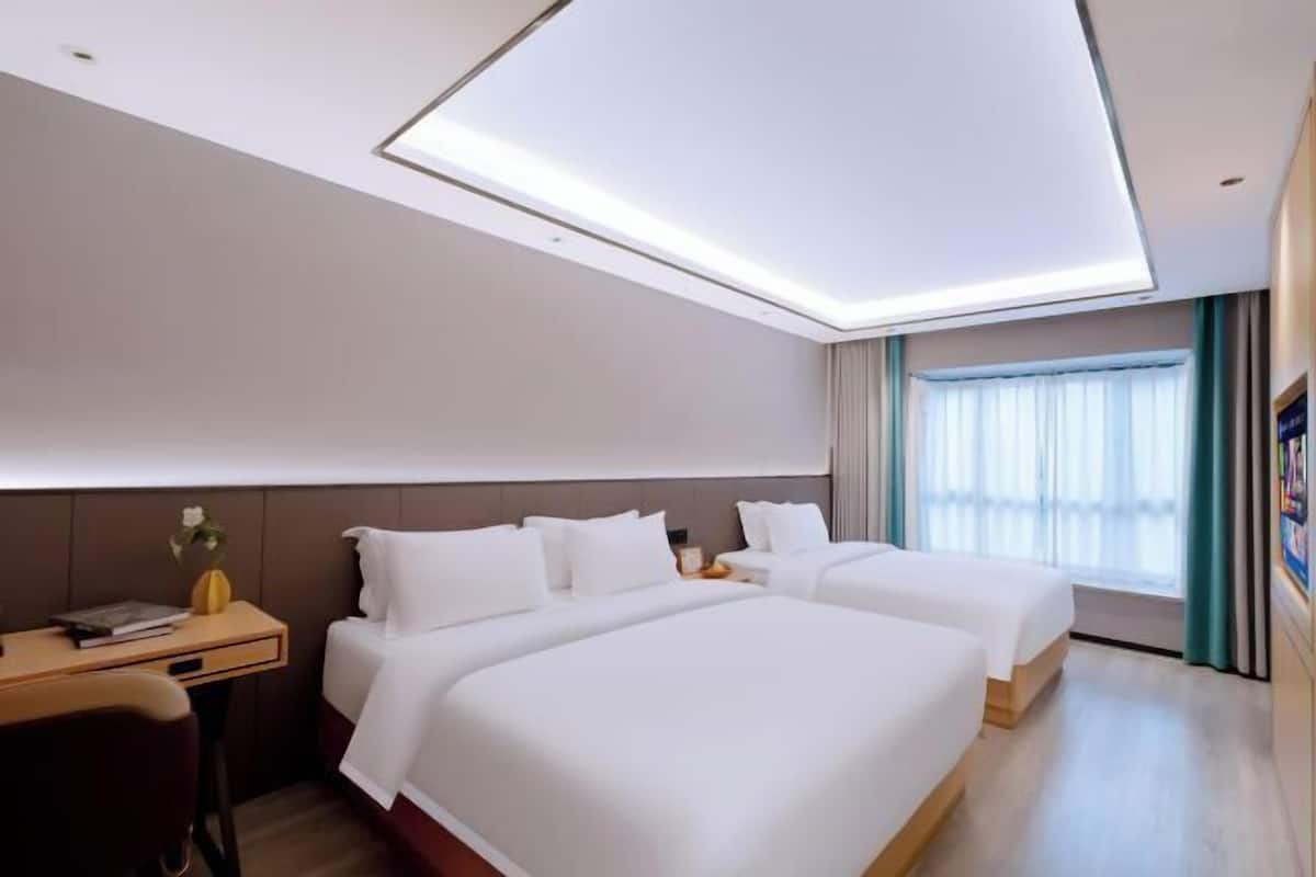 Hotellihuone, jossa on kaksi sänkyä, työpöytä, maljakko kukkineen ja suuri ikkuna, jossa on verhot.