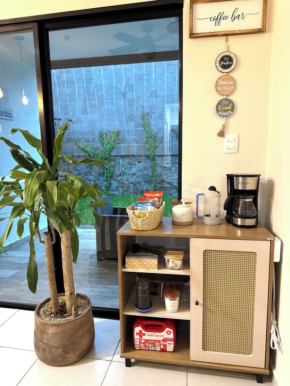 Un rincón acogedor con cafetería, una planta en maceta, una cesta de aperitivos, una máquina de café y un botiquín.