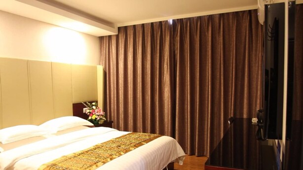 Habitación de hotel con cama, televisor, mesita de noche con un florero y un ventanal con cortinas.