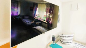 Una habitación con cama, ventana con cortinas, una mesita de noche con lámpara y televisor instalado en la pared.