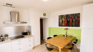 Una cocina moderna con comedor, armarios blancos, campana extractora de acero inoxidable, cocina de inducción negra, una mesa de madera con cuatro sillas y un cuadro colorido en la pared.