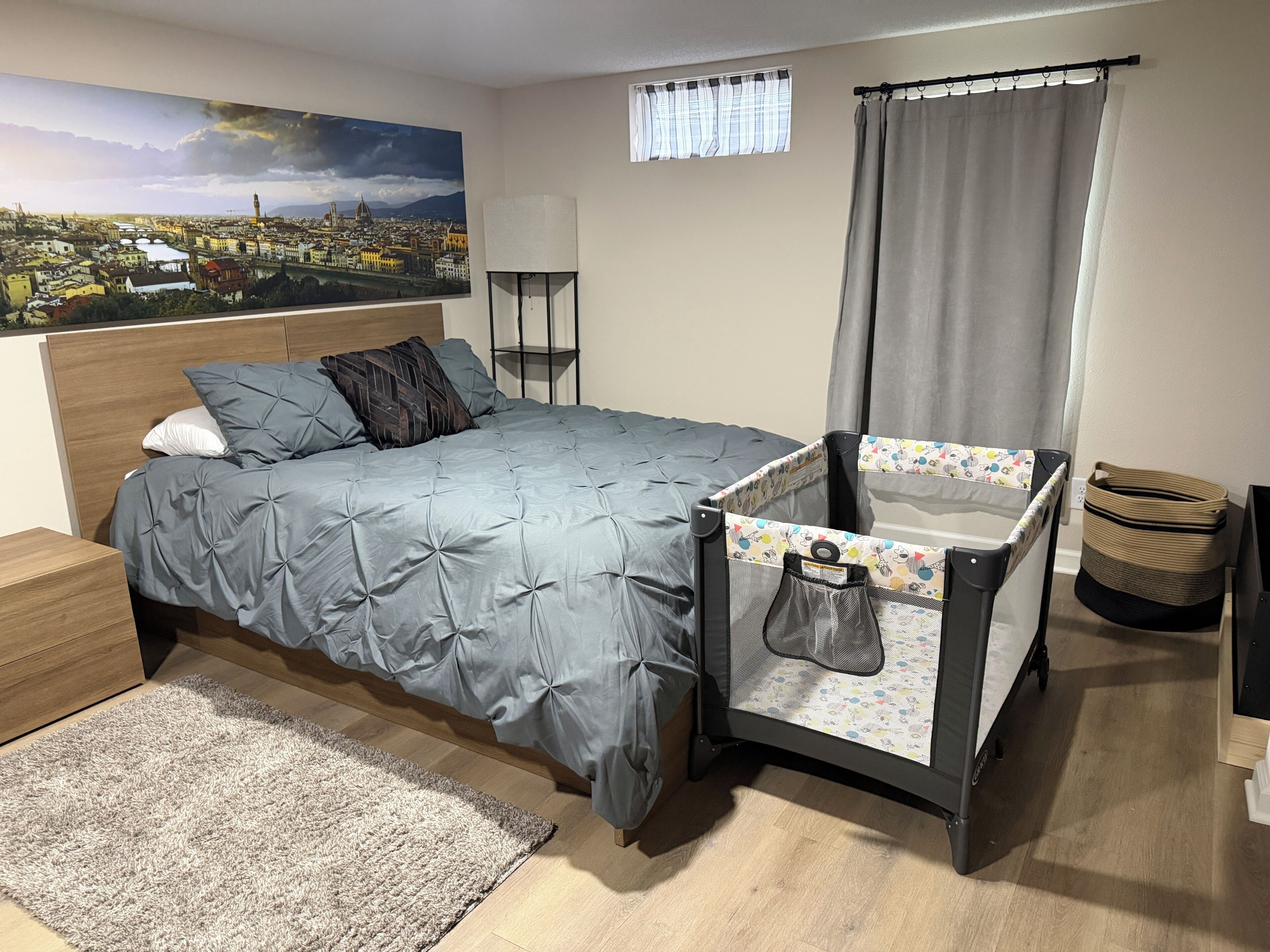 Ein Schlafzimmer mit einem Bett, einem Babybett, einem Nachttisch, einer Lampe, einem Fenster mit Vorhängen und einem Stadtbild an der Wand.