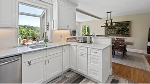 Una cocina moderna con armarios blancos, un ventanal grande y una zona de comedor con mesa y sillas de madera.
