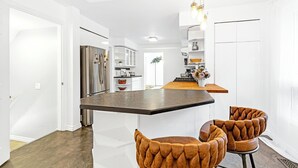 A modern kitchen with white cabinetry, a stainless steel refrigerator, and a bar area with two brown upholstered chairs. - Luxury Winery Country Retreat: Heated Pool, Hot Tub and Games (Niagara-on-the-Lake)
